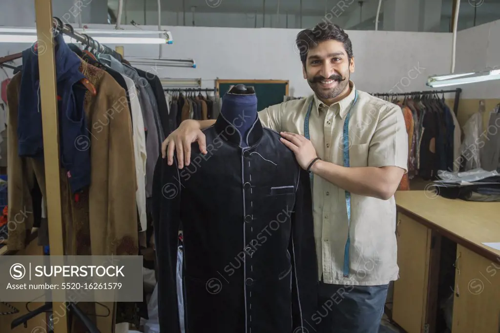 Tailor standing beside a mannequin wearing a semi-stitched coat in his workshop.