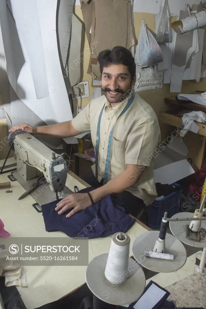 Happy looking tailor working on sewing machine stitching a cloth in his small workshop.