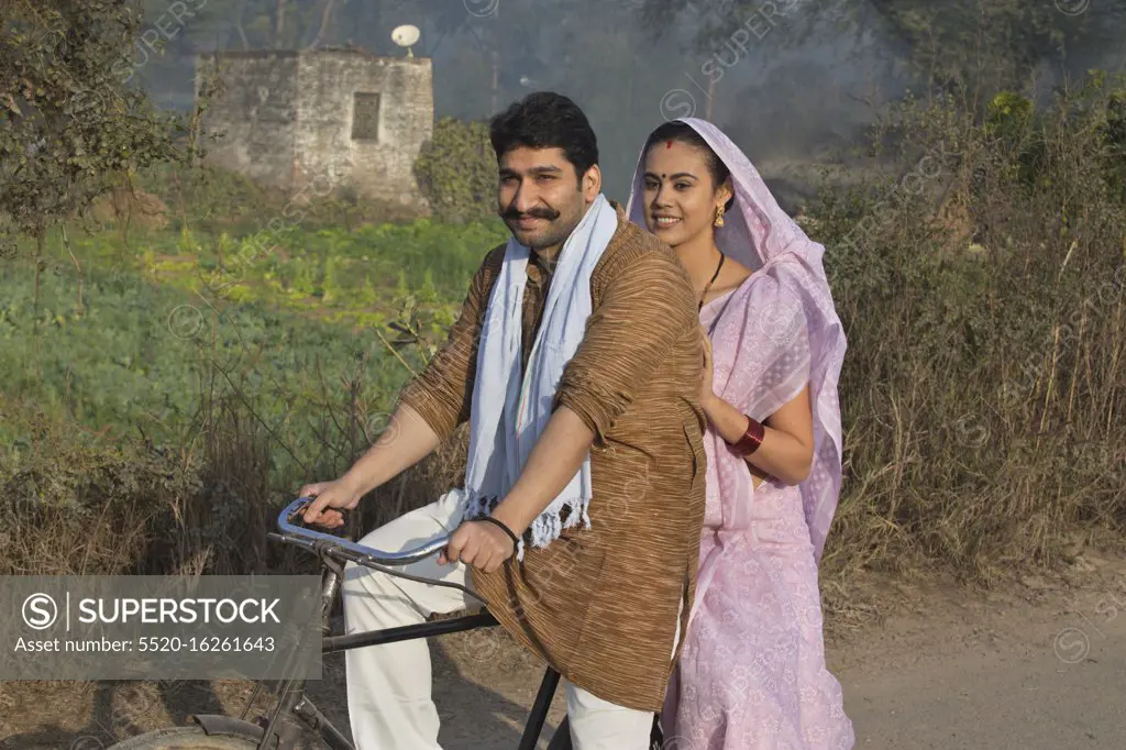 Side view of a happy rural couple in traditional dress riding on bicycle in a village.