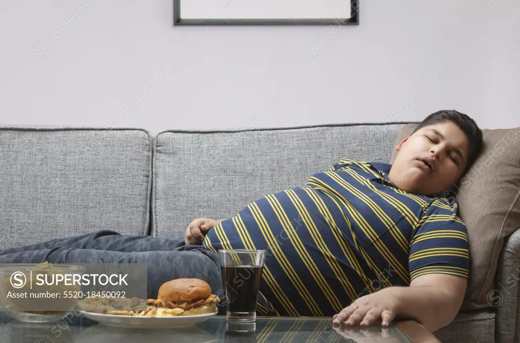 Young boy sleeping on the sofa with junk food kept on the table  at home. (Obesity) 