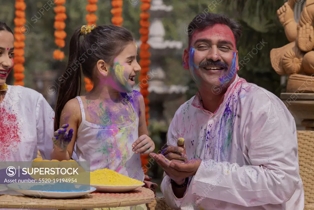 Granddaughter applying colour on grandfather's face and mother sitting beside on the occasion of Holi