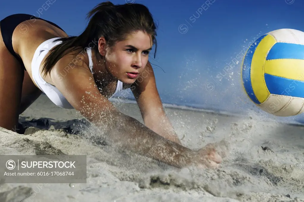 Young woman lunging onto sand to hit a volleyball upwards before it hits the ground