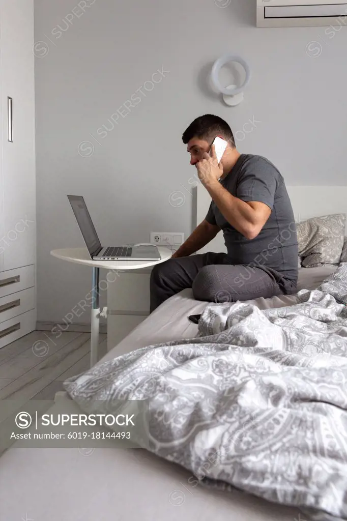 Man talking on the phone, working behind a laptop in the bedroom on th