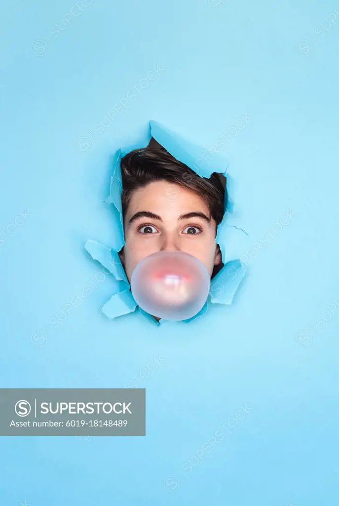 Boy blowing bubble with head through hole in blue paper background.