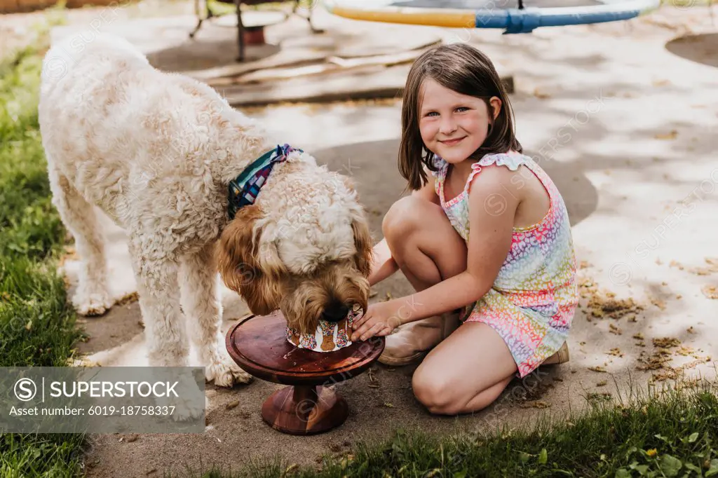 Young girl holding cake while dog eats it outside in backyard