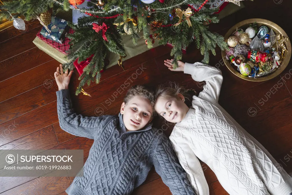 Brother and sister are lying on the floor under the Christmas tree.