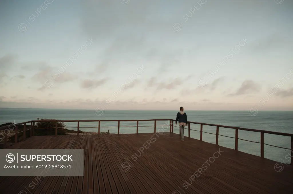 Woman walking on boardwalk against ocean at sunset in Sagres, Portugal