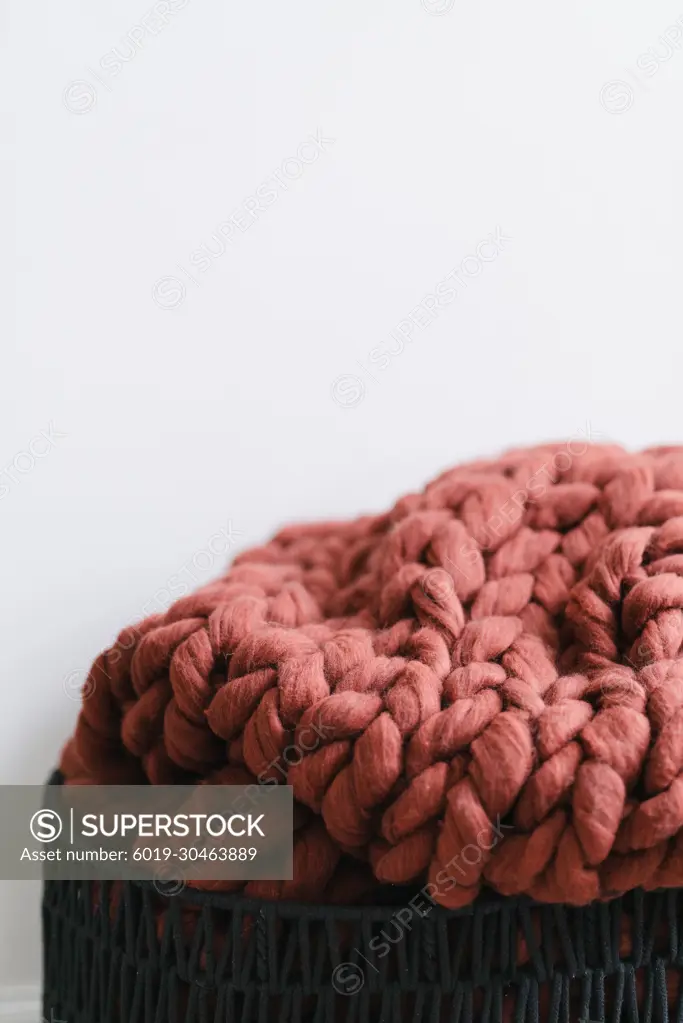 Rust chunky knit blanket in black basket by white wall SuperStock