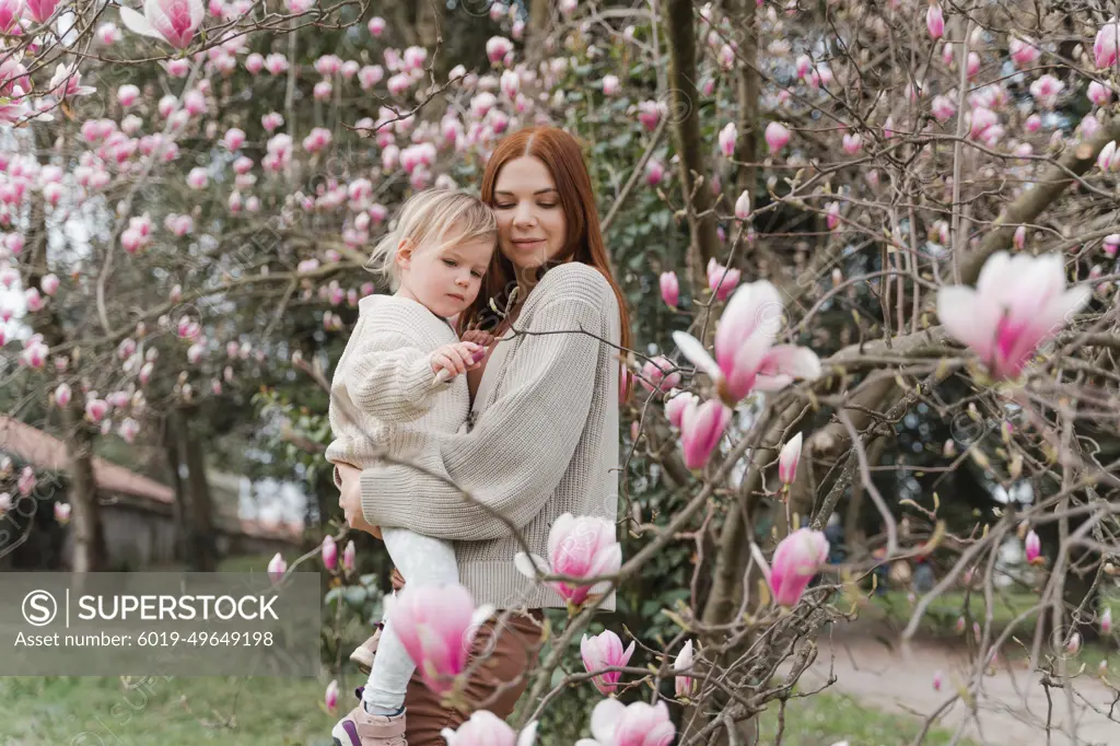 A woman hugs her little daughter near a blooming magnolia tree.