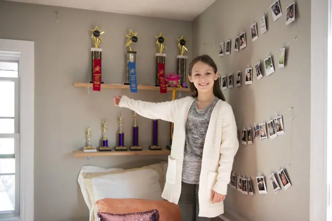Young girl standing on her bed by a shelf of dance trophies.