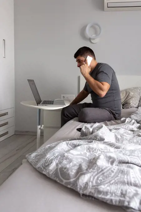 Man talking on the phone, working behind a laptop in the bedroom on th