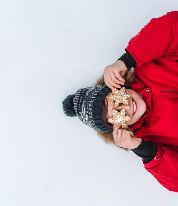 Overhead of boy with snowflake cookies over eyes laying in the snow.