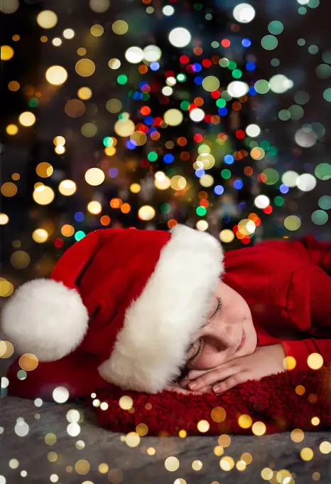 Portrait of boy wearing Santa hat sleeping in front of Christmas tree.