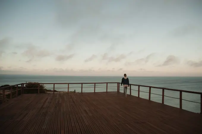 Woman walking on boardwalk against ocean at sunset in Sagres, Portugal