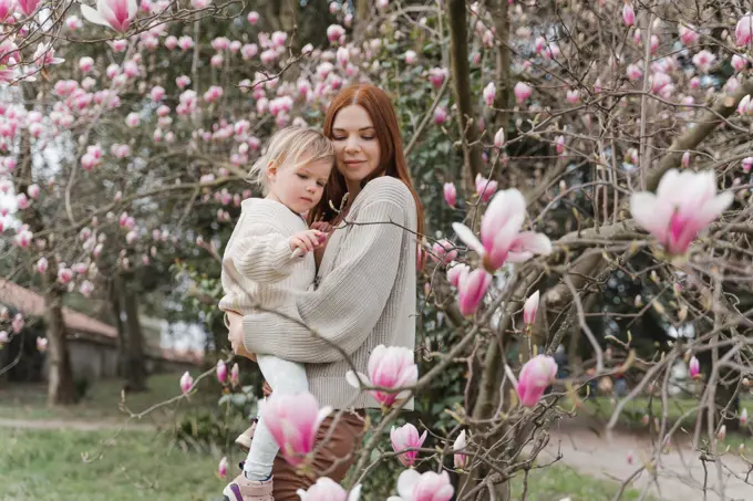 A woman hugs her little daughter near a blooming magnolia tree.