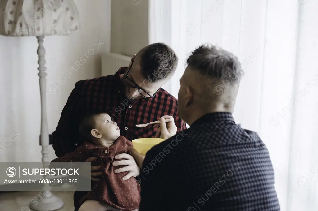 Male gay couple with adopted baby girl at home - Two handsome dads feed the baby girl on kitchen - Male babysitters - Lgbt family at home - Diversity concept