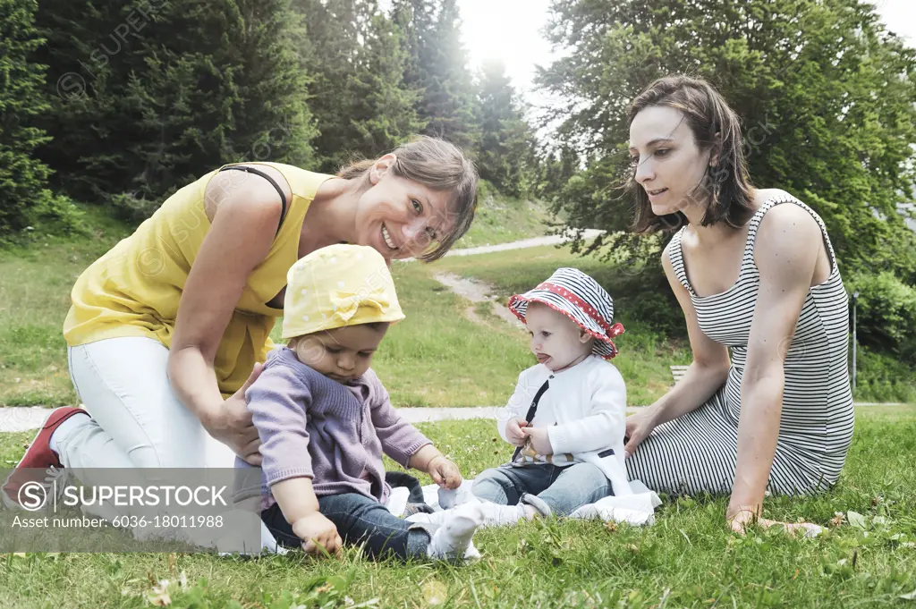 Two friend women enjoying a picnic in a beautiful sunny day with their little daughters - Women sitting with little baby girls on grass in the mountain park - Concept of motherhood and real friendship