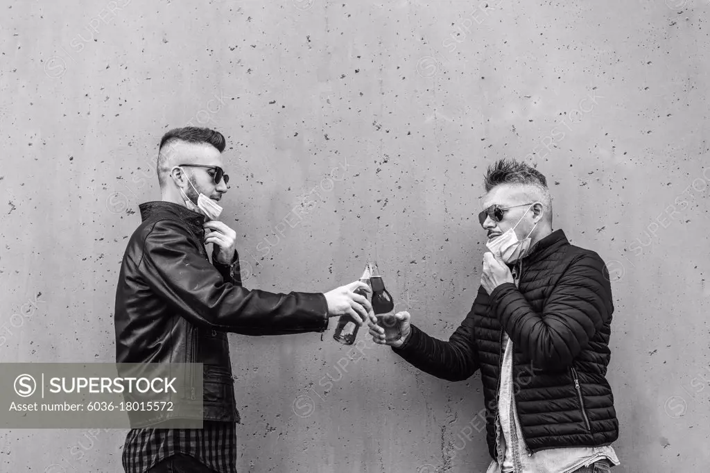 Guys friends toasting beer bottles with opened face masks - New normal lifestyle concept with people having fun together drinking but with social distancing - Black and white version with copy space