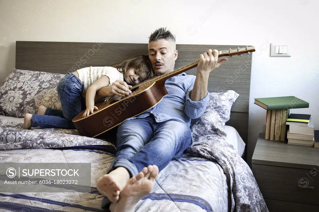 Cool daddy playing the acoustic guitar and teaching a song while his daughter listens to him - Dad and daughter are sitting on the bed playing guitar - Teaching and parenthood concept  - Father's Day