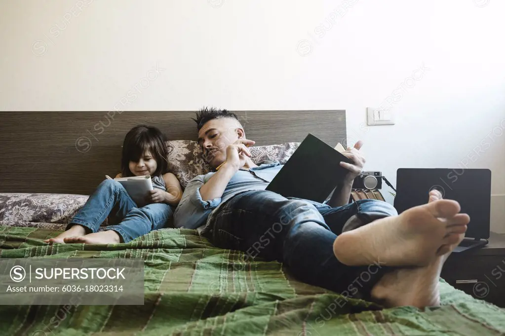 Father and daughter in casual denim clothes are sitting on bed together. The girl having fun writing or drawing while the father looks her. Childhood and parenthood concept. Leisure activity at home.