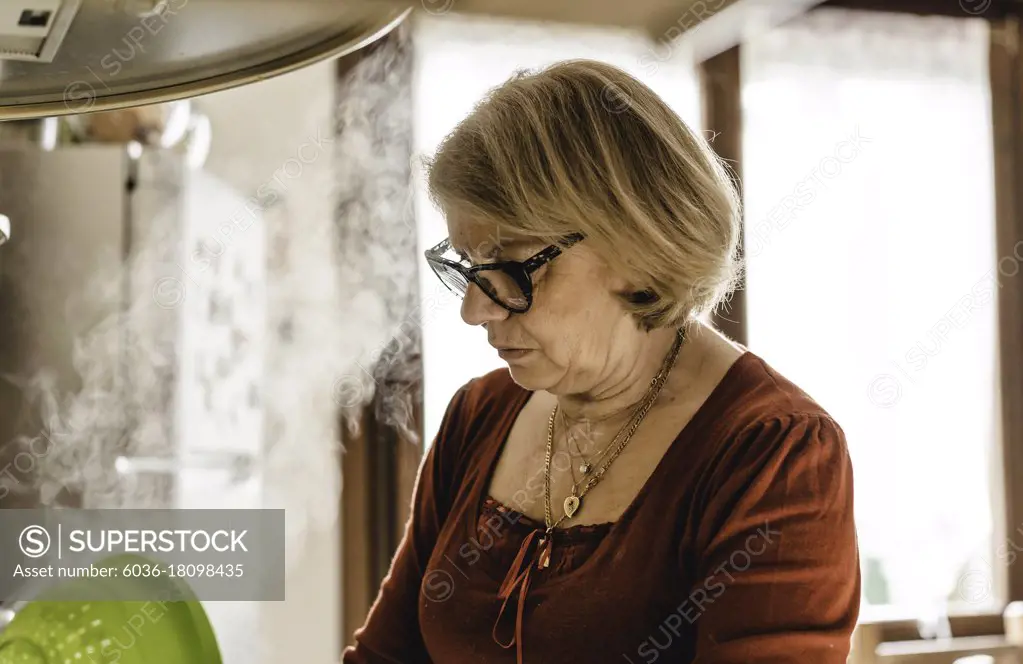 Senior woman cooking lunch in the kitchen - Mature woman in the kitchen draining the pasta from the boiling water -  Boomer woman busy cooking lunch for the family