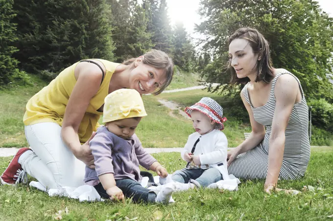 Two friend women enjoying a picnic in a beautiful sunny day with their little daughters - Women sitting with little baby girls on grass in the mountain park - Concept of motherhood and real friendship