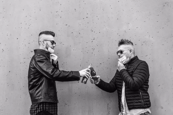 Guys friends toasting beer bottles with opened face masks - New normal lifestyle concept with people having fun together drinking but with social distancing - Black and white version with copy space