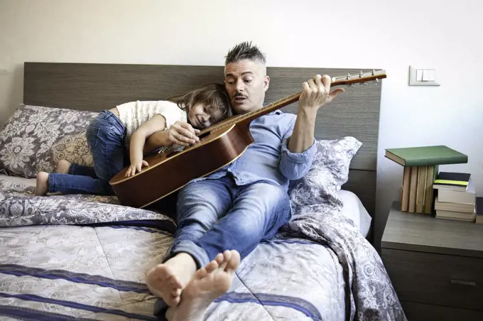 Cool daddy playing the acoustic guitar and teaching a song while his daughter listens to him - Dad and daughter are sitting on the bed playing guitar - Teaching and parenthood concept  - Father's Day