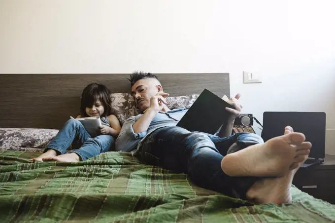 Father and daughter in casual denim clothes are sitting on bed together. The girl having fun writing or drawing while the father looks her. Childhood and parenthood concept. Leisure activity at home.