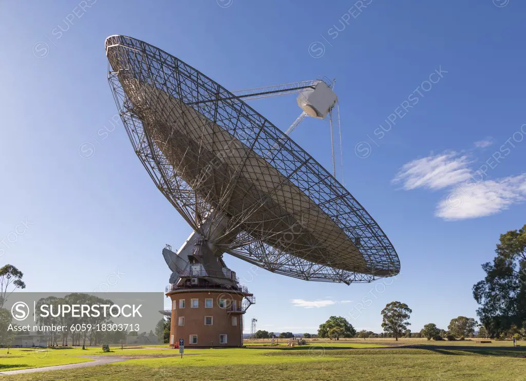 A large outdoor scientific radio telescope in the sunshine in a green field with trees