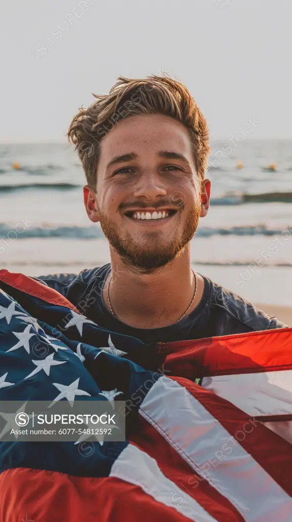 A man standing on the beach, proudly holding an American flag. The ocean waves in the background, under a clear sky.