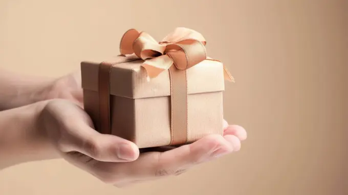 A pair of hands hold out a small, square gift box tied with a brown satin ribbon and bow. The gift box is wrapped in brown paper and the background is a light brown color.