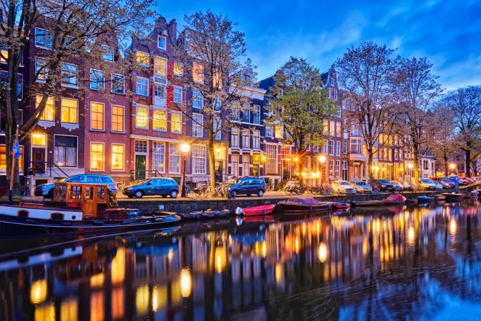 Night view of Amsterdam cityscape with canal, boats and medieval houses in the evening twilight illuminated. Amsterdam, Netherlands