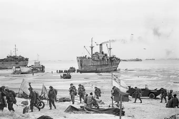 Troops come ashore on one of the Normandy invasion beaches, past the White Ensign of a naval beach party, 7 June 1944. D-Day, 6 June 1944: The White Ensign of a Naval Beach Party flying on the Normandy Coast as British troops move inland from the beached landing craft.