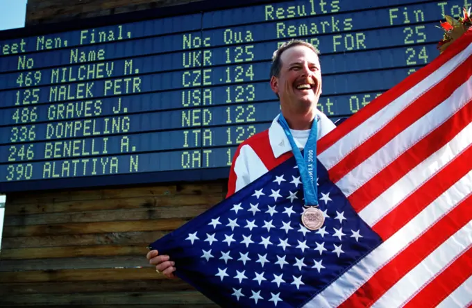 Straight on, medium shot from a low angle, as US Army Sergeant First Class James Graves stands holding an American flag, wears a Bronze medal around his neck and is in front of a large scoreboard. SFC Graves, from the US Army Marksmanship Unit at Fort Benning, Georgia, won his Bronze Medal on September 23rd, 2000, in the Men's Skeet competition at the 2000 Olympic games in Sydney, Australia. Base: Sydney State: New South Wales Country: Australia (AUS)