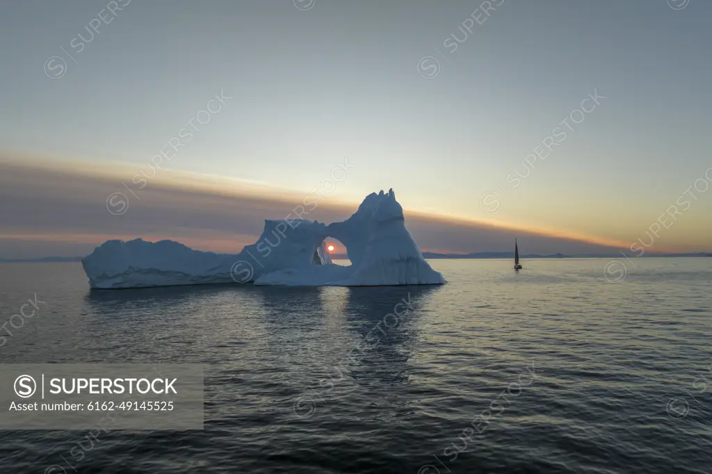 Aerial drone view of the sunset through a huge iceberg arch surrounded by a sailing boat, Ilulissat, Greenland, Arctic.