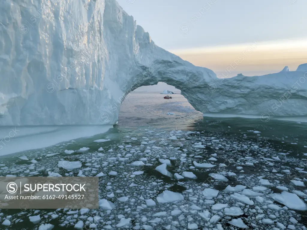 Aerial drone view of a red boat though a huge iceberg arch, Ilulissat, Greenland, Arctic.