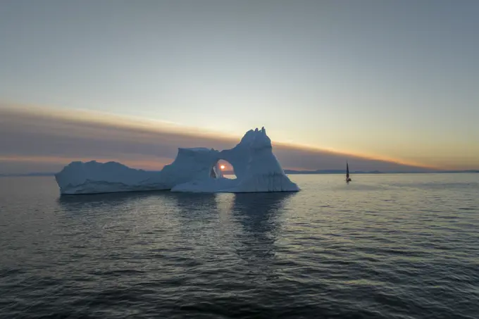 Aerial drone view of the sunset through a huge iceberg arch surrounded by a sailing boat, Ilulissat, Greenland, Arctic.