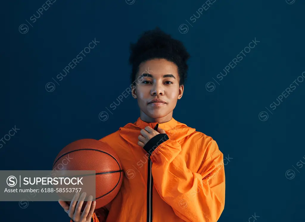 Young female adjusting her orange sportswear and looking at the camera over a blue backdrop. Professional female basketball player with the ball in the studio. Young female adjusting her orange sportswear and looking at the camera over a blue backdrop. Professional female basketball player with the ball in the studio. Copyright: xxArtemxVarnitsinx