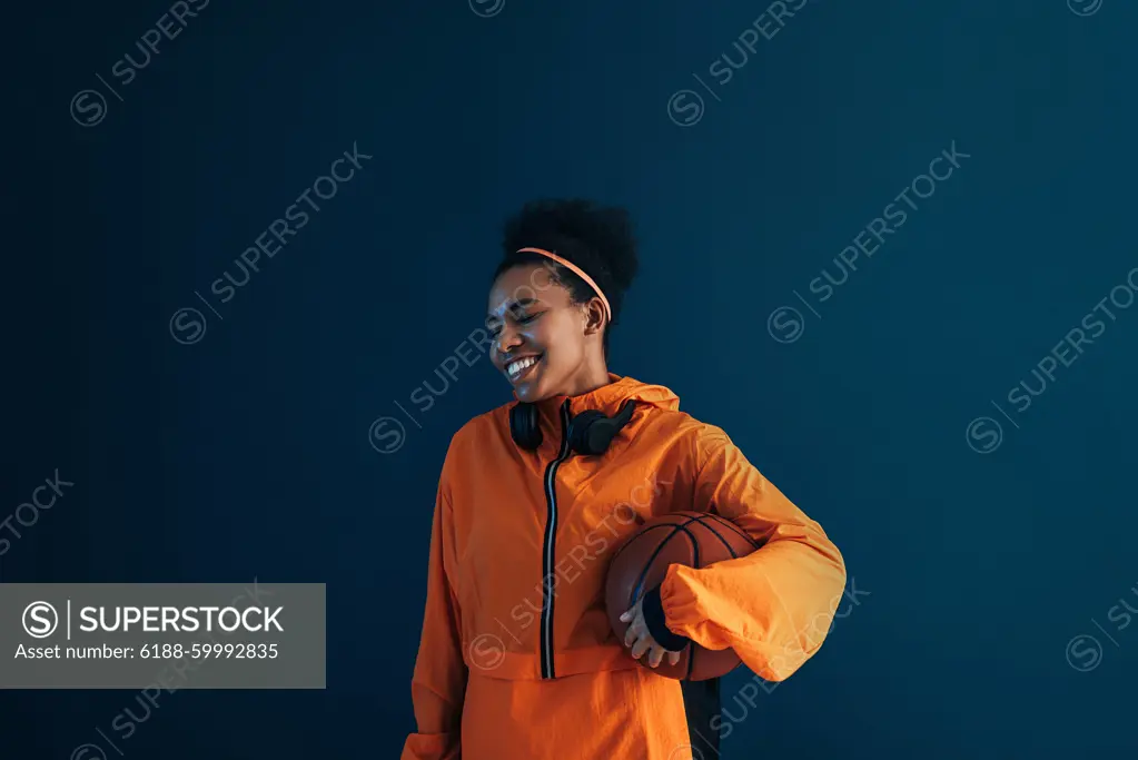 Cheerful female in orange sports attire over blue backdrop. Young smiling professional basketball player in the studio. Cheerful female in orange sports attire over blue backdrop. Young smiling professional basketball player in the studio. Copyright: xxArtemxVarnitsinx