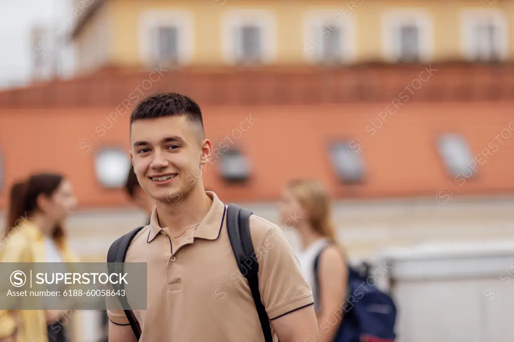 Close-up of a male portrait of a cute guy in a beige T-shirt with a black backpack. The teenager smiles and looks at the camera. Close-up of a male portrait of a cute guy in a beige T-shirt with a black backpack. The teenager smiles and looks at the camera. Roofs of houses and other teens in the background. ,model released, Symbolfoto Copyright: xZoonar.com/DashaxPetrenkox 21660161 ,model released, Symbolfoto ,property released