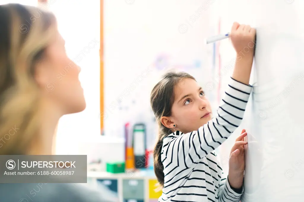 Hardworking teacher looking at young schoolgirl in classroom. Girl student writing on whiteboard. Hardworking teacher looking at young schoolgirl in classroom. Girl student writing on whiteboard with marker. model released