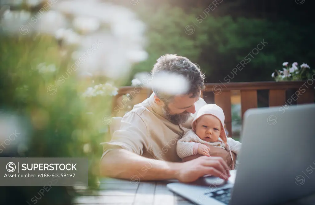 Dad working from homeoffice, sitting on patio with laptop, holding little baby girl. Father spending time with small daughter, enjoying together time. Father s Day. Dad working from homeoffice, sitting on patio with laptop, holding little baby girl. Father spending time with small daughter, enjoying together time. Father s Day concept. model released