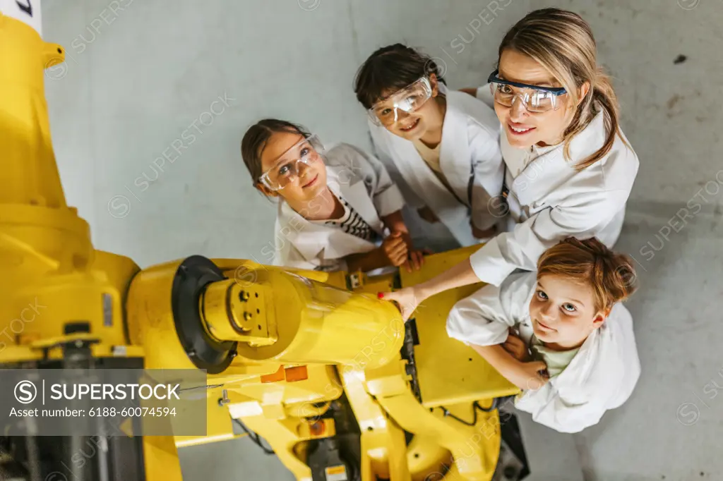 Top view of children learning robotics in Elementary school, standing with female teacher by robotic arm. After-school robotics club, or field trip to real robotics laboratory. Portrait of children learning robotics in Elementary school, standing with female teacher. Young students building robot in after school robotics club. Field trip to real robotics laboratory. model released