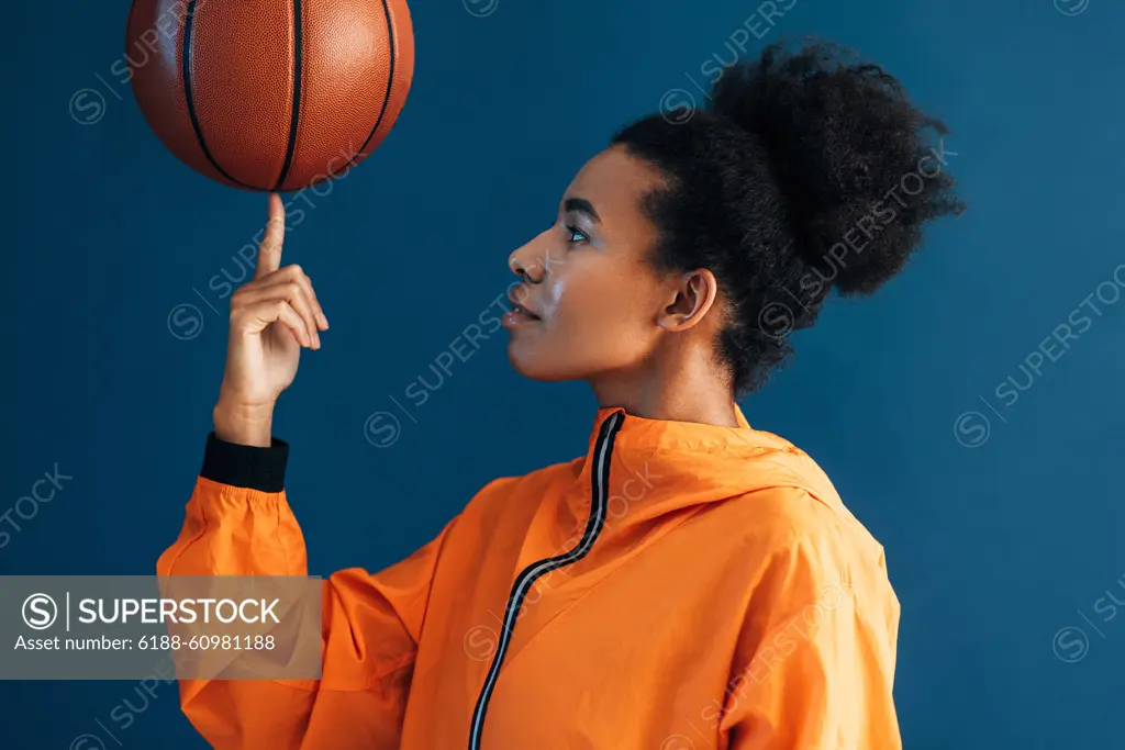 Side view of young woman with curly hair holding basketball on finger. Studio shot of female basketball player posing in studio. Side view of young woman with curly hair holding basketball on finger. Studio shot of female basketball player posing in studio. Copyright: xxArtemxVarnitsinx