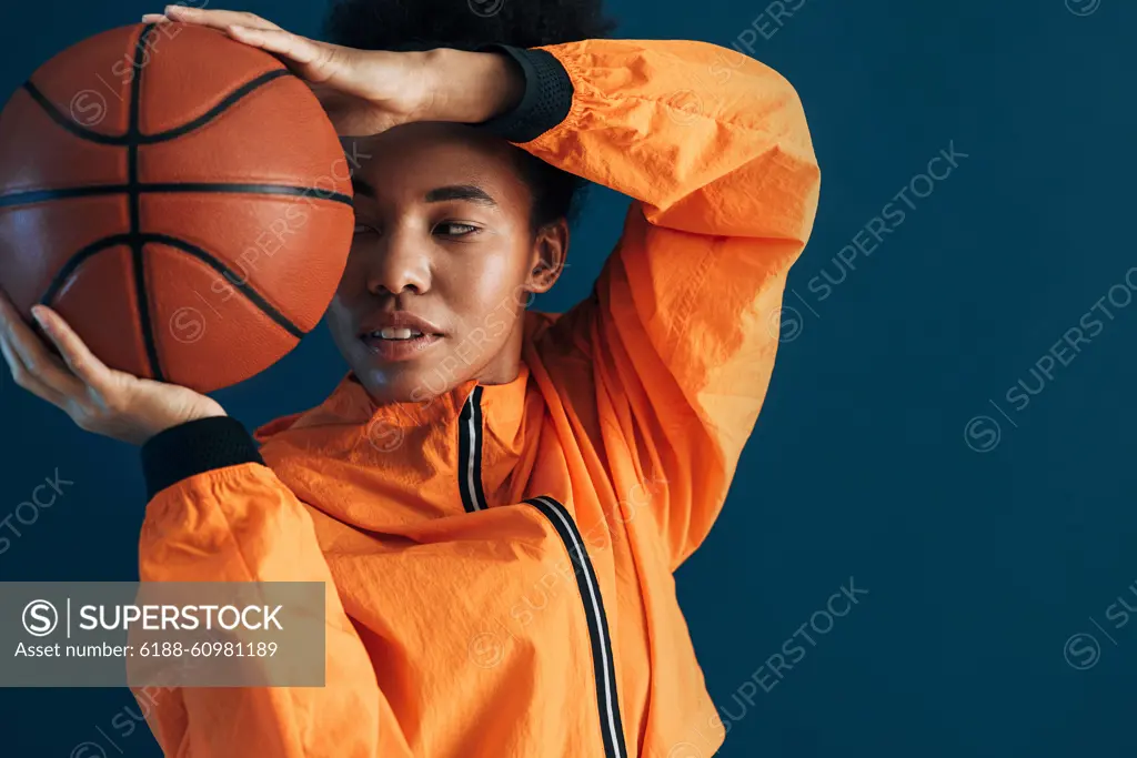 Close up studio shot of young female basketball player in orange sportswear posing with ball. Portrait of a professional basketball player over blue background. Close up studio shot of young female basketball player in orange sportswear posing with ball. Portrait of a professional basketball player over blue background. Copyright: xxArtemxVarnitsinx