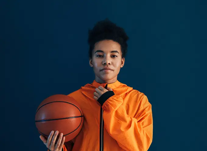 Young female adjusting her orange sportswear and looking at the camera over a blue backdrop. Professional female basketball player with the ball in the studio. Young female adjusting her orange sportswear and looking at the camera over a blue backdrop. Professional female basketball player with the ball in the studio. Copyright: xxArtemxVarnitsinx