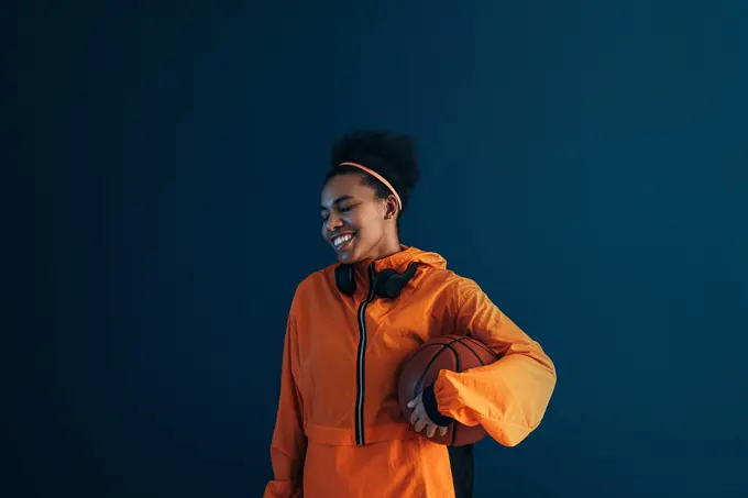 Cheerful female in orange sports attire over blue backdrop. Young smiling professional basketball player in the studio. Cheerful female in orange sports attire over blue backdrop. Young smiling professional basketball player in the studio. Copyright: xxArtemxVarnitsinx