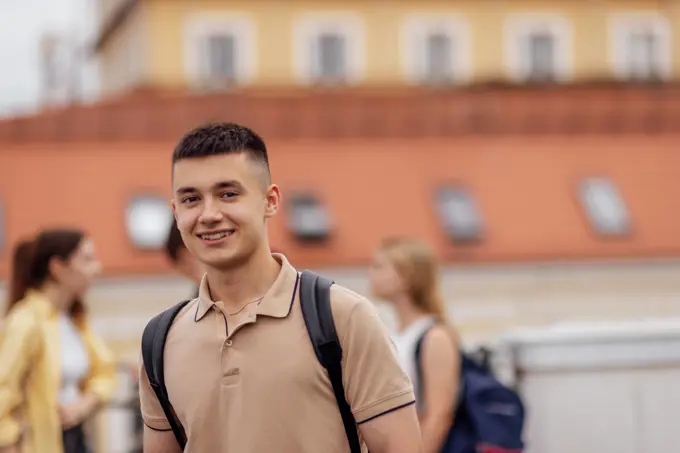 Close-up of a male portrait of a cute guy in a beige T-shirt with a black backpack. The teenager smiles and looks at the camera. Close-up of a male portrait of a cute guy in a beige T-shirt with a black backpack. The teenager smiles and looks at the camera. Roofs of houses and other teens in the background. ,model released, Symbolfoto Copyright: xZoonar.com/DashaxPetrenkox 21660161 ,model released, Symbolfoto ,property released