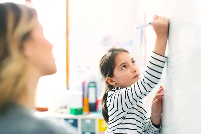 Hardworking teacher looking at young schoolgirl in classroom. Girl student writing on whiteboard. Hardworking teacher looking at young schoolgirl in classroom. Girl student writing on whiteboard with marker. model released